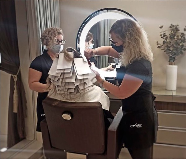 Imagem com pessoas tingindo o cabelo de uma cliente. Nessa imagem, as funcionárias estão de máscara sob um anel de luz.