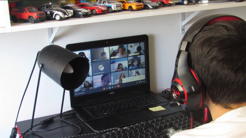 #ParaTodosVerem: Criança utilizando fone, sentada em frente ao computador durante aula online. Na tela do computador podem ser observados outros colegas de sala.