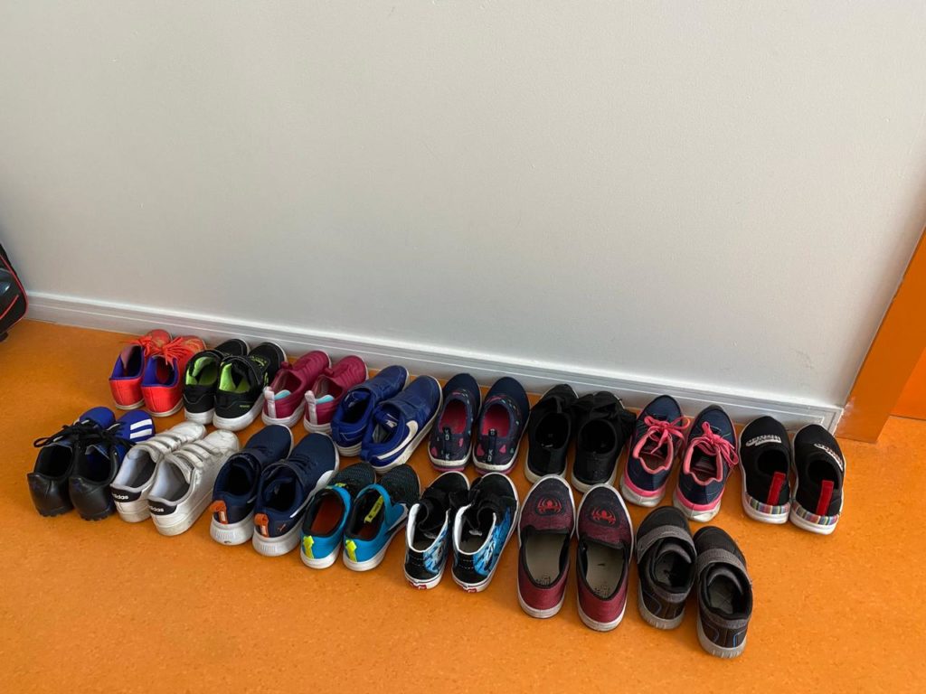 #ParaTodosVerem: sapatos infantis enfileirados no chão laranja de uma sala de aula. As crianças devem retirar os sapatos antes de entrar na classe.