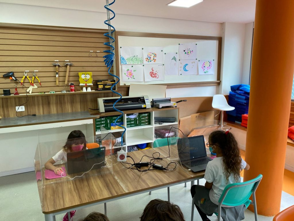 #ParaTodosVerem: Duas crianças sentadas olhando para o computador enquanto estudam. Separadas por placas de acrílico e utilizando máscaras. 