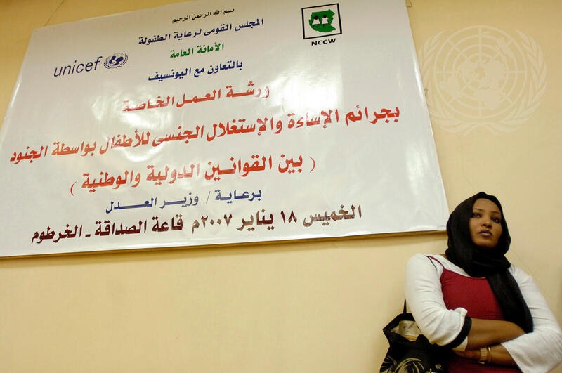 Na imagem: mulher sudanesa em frente a um banner que anuncia um workshop sobre exploração sexual e abuso