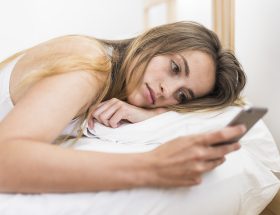 Foto ilustrativa de uma mulher com celular e deitada na cama